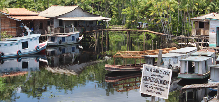 A fotografia apresenta uma visão geral da Comunidade Santo Ezequiel Moreno, Portel/PA. O primeiro plano mostra, no canto inferior direito, uma placa com informações da localização da Comunidade. No segundo plano, à direita, dois barcos brancos e um terceiro, ainda em construção, estão ancorados. À esquerda, dois outros barcos estão ancorados na frente de duas casas de palafita. No terceiro plano, uma vegetação verde e densa compõe a imagem. Está de dia.